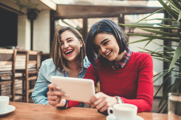 Two Young Woman Holding tablet, Chatting and smiling in a Coffee Shop.