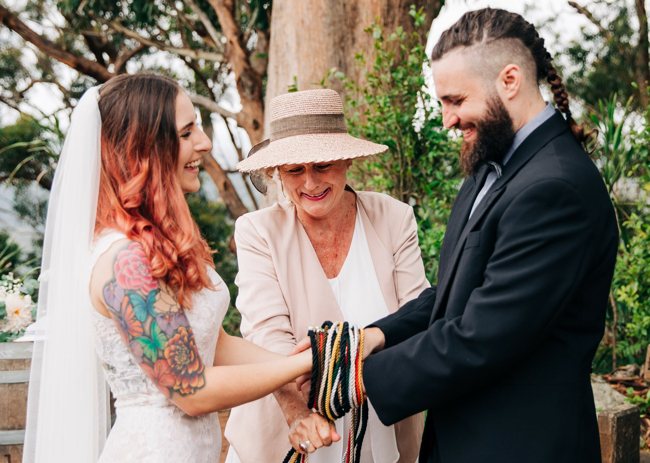 A bride with bright tattoos and a grown with a Viking braid clasp hands as their celebrant winds coloured cords around their hands.